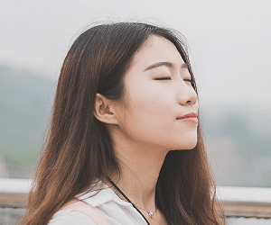 A young woman stands outside with her eyes closed and a relaxed expression.