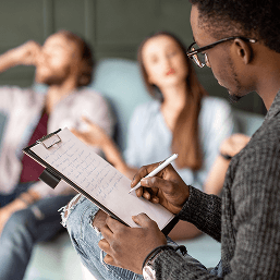 A male therapist wearing glasses  takes notes on a clipboard as he counsels a young couple sitting on a couch