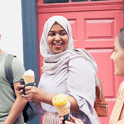 Three smiling college students wearing backpacks are walking down the street eating ice cream cones