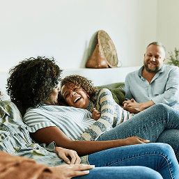 Two relaxed women laugh and hug on a couch along with a smiling male family member sitting at the end
