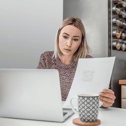 Alt text: A stressed looking blonde woman reads a sheet of paper while working with a laptop