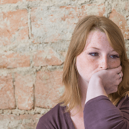 A stressed young woman sits against a wall looking away with her hand over her mouth