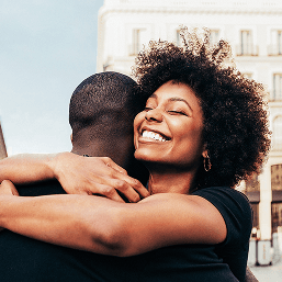 A young woman smiles brightly as she embraces a man on a sunny day