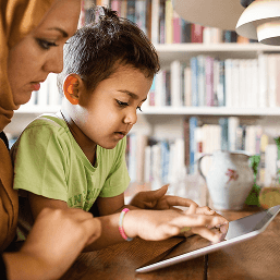In a library, a mother wearing a hijab looks on as her son does an activity on a tablet